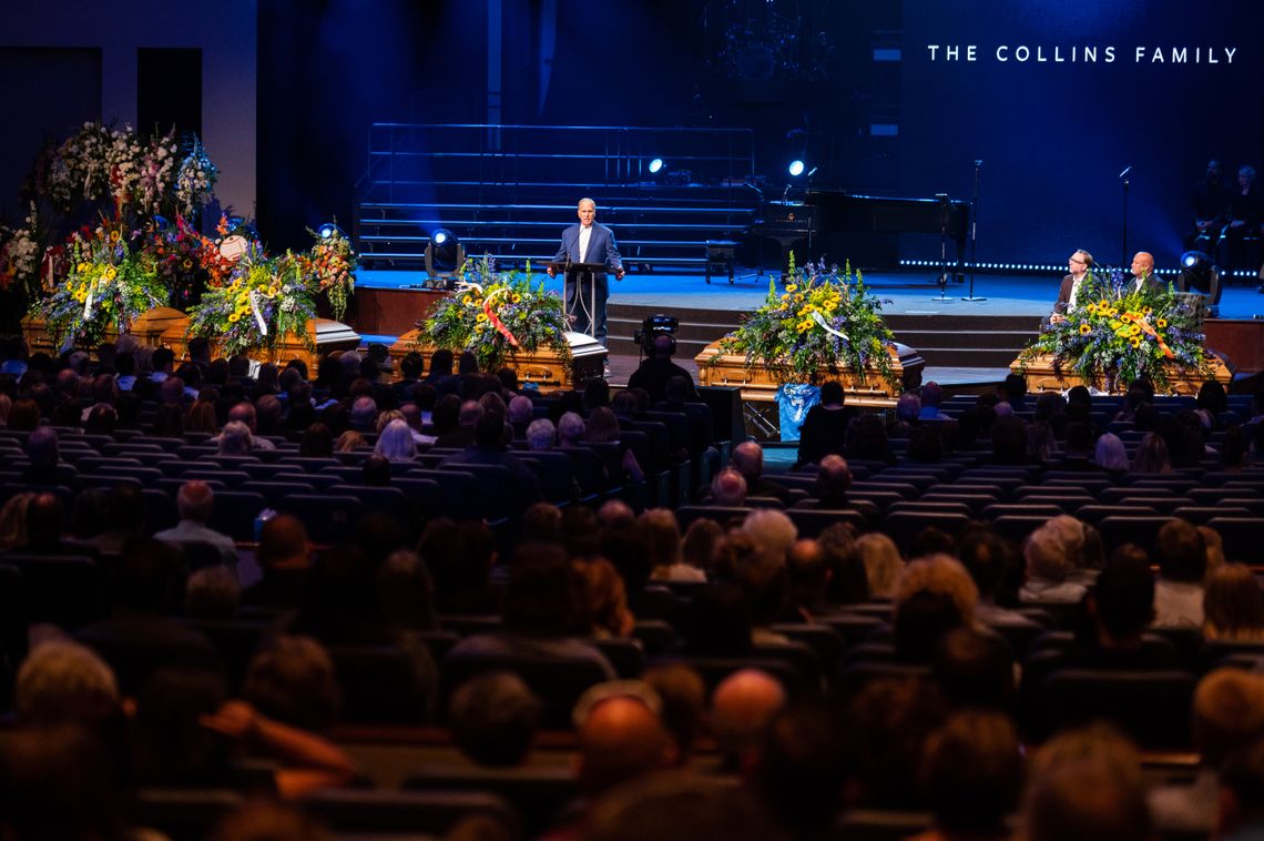 Five coffins adorned with flowers are situated in front of a White man speaking at a podium of a large church. 