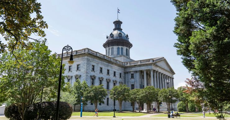 A photograph from an angle of the South Carolina State House, a light-colored building with a triangular pediment and columns in front, and trees on a lawn with paths. 
