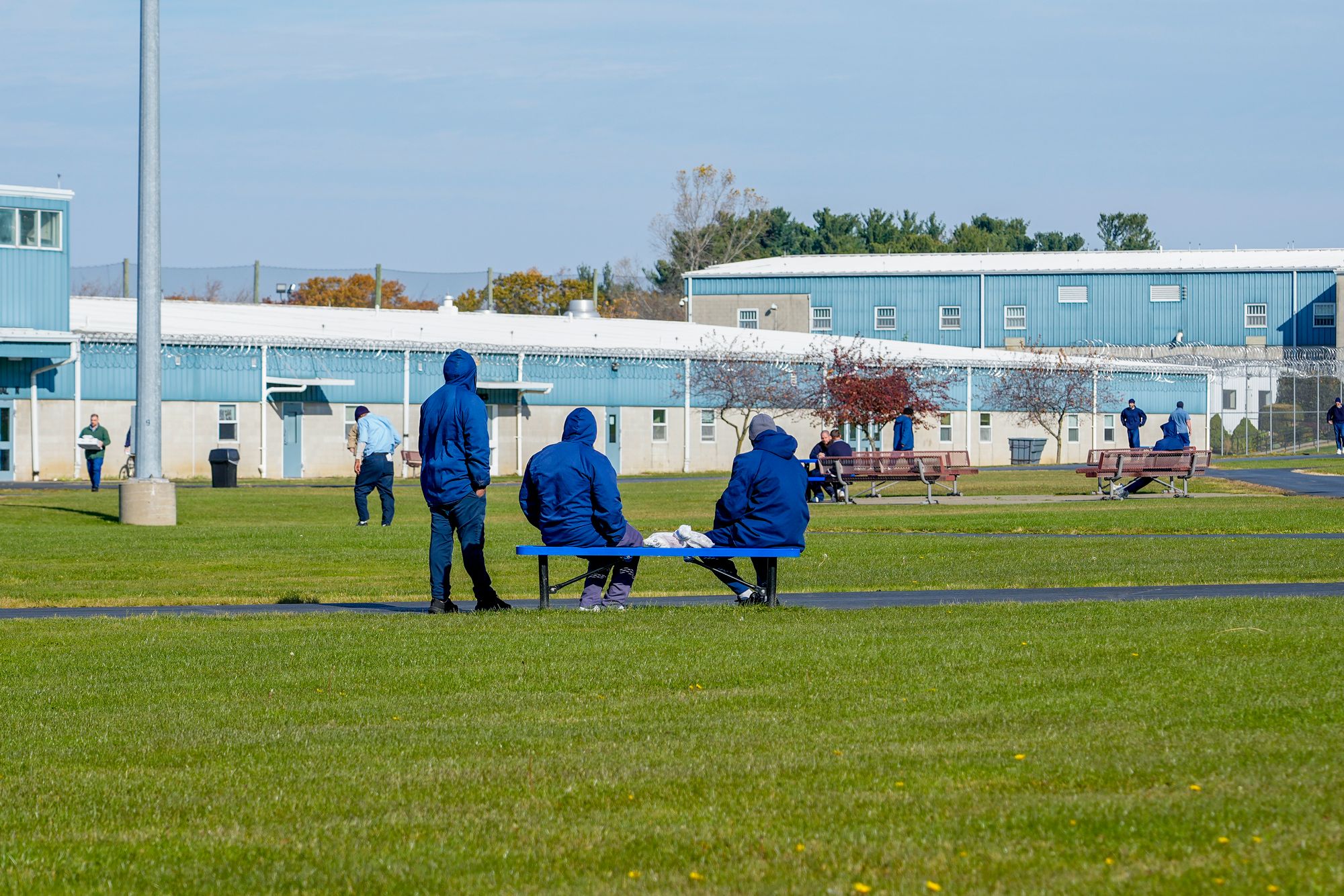A photo shows three incarcerated people dressed in blue uniforms in the foreground, outdoors, but on prison grounds. Their faces are not visible, and two of them are sitting on a bench, and one is standing to the left. Two buildings are visible behind the three men, which are pale blue and white, and there are a few other people visible in the background. In the distance, netting along the fence line is visible. 
