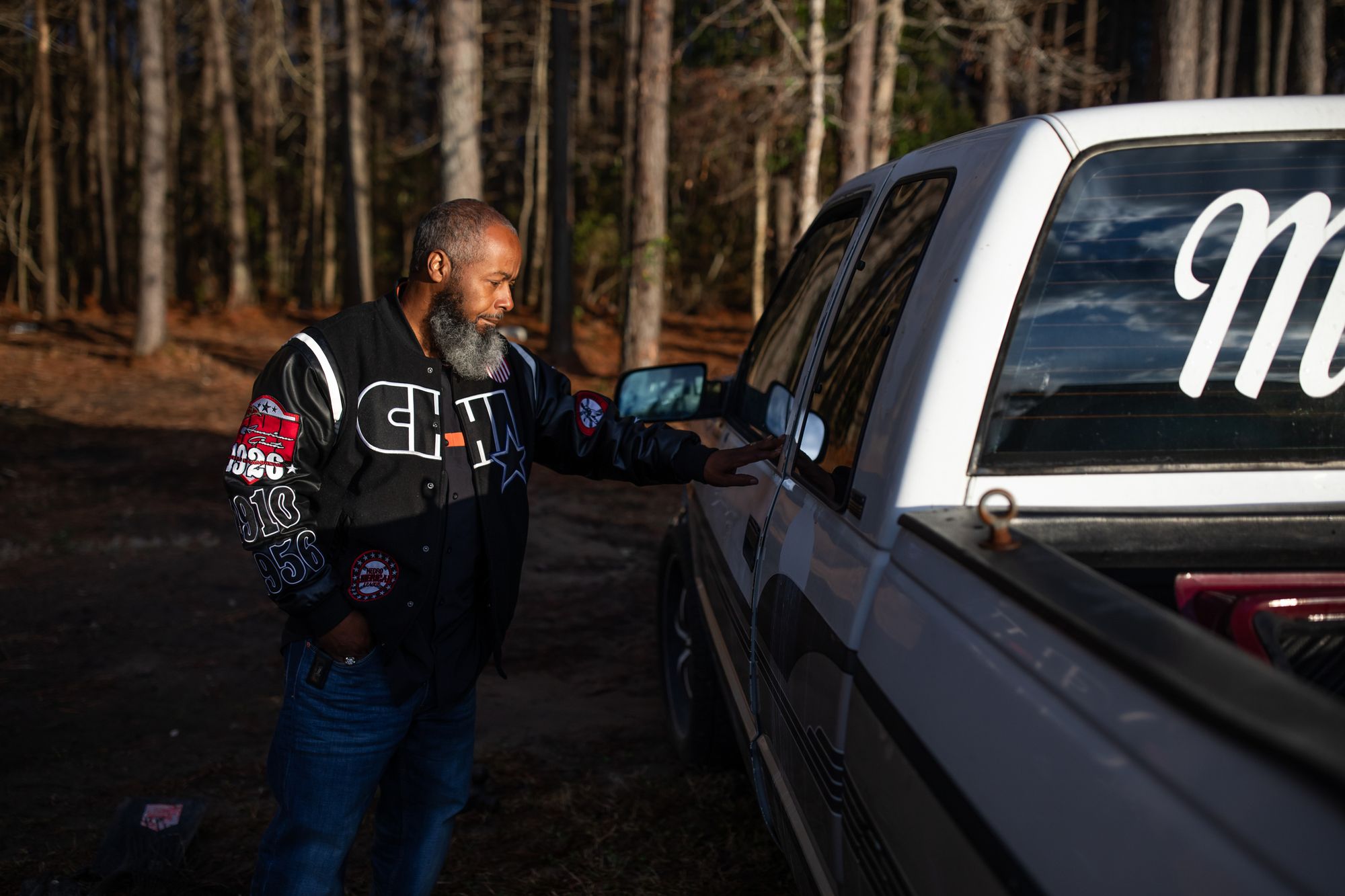 A photo shows a Black man with a beard and black patterned jacket standing next to a white truck. He is touching the weather stripping of the window with his hand. 