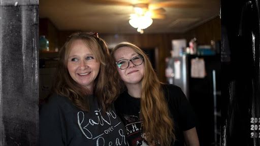 A White woman with brown hair and blond highlights leans against a wall, while her daughter, a White woman with glasses and long, brown-blond hair, leans into her.