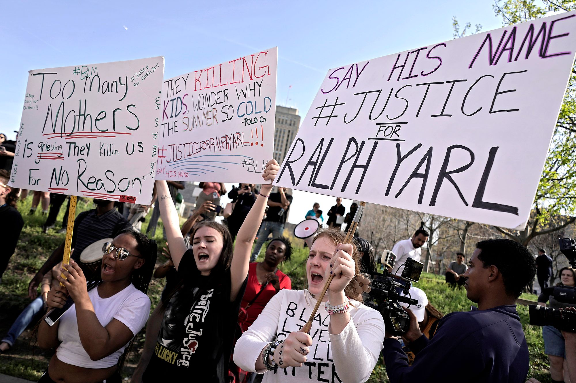Protesters at a park hold signs that reads "Too many mothers is grieving, they killing us for no reason," "They killing kids, wondering why the summer so cold. Justice for Ralph Yarl!!" and "Say his name, justice for Ralph Yarl."  People with bullhorns and cameras stand in the background. 
