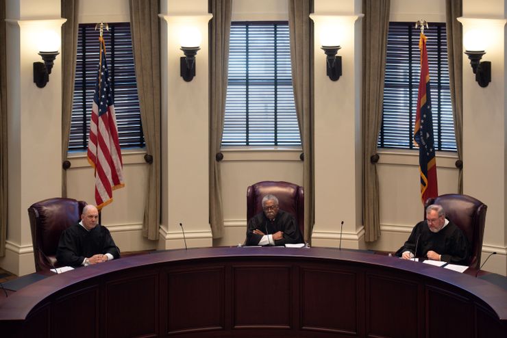 A photo shows three men in black robes sitting at a large, curved judges’ bench in a courtroom. Two of the men are White, and one, in the center, is Black.  