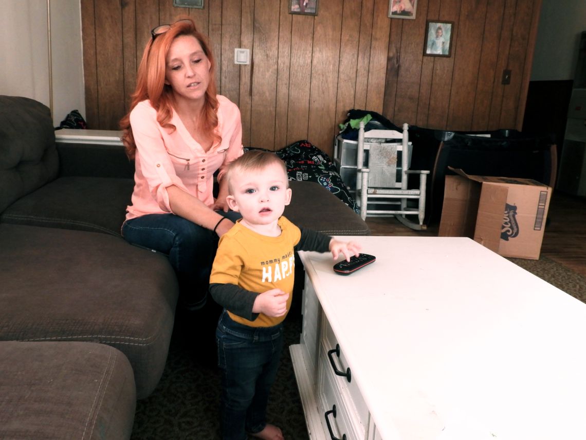 A woman with light-toned skin and reddish long hair sits on a sofa, with a toddler in front of her.  