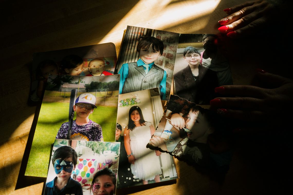 Sunlight falls on a group of seven photos on a table showing pictures of Rebecca McDonald's son. There are photos of him as a baby, a toddler, and a young boy. McDonald's hands rest on the photos on the right. 