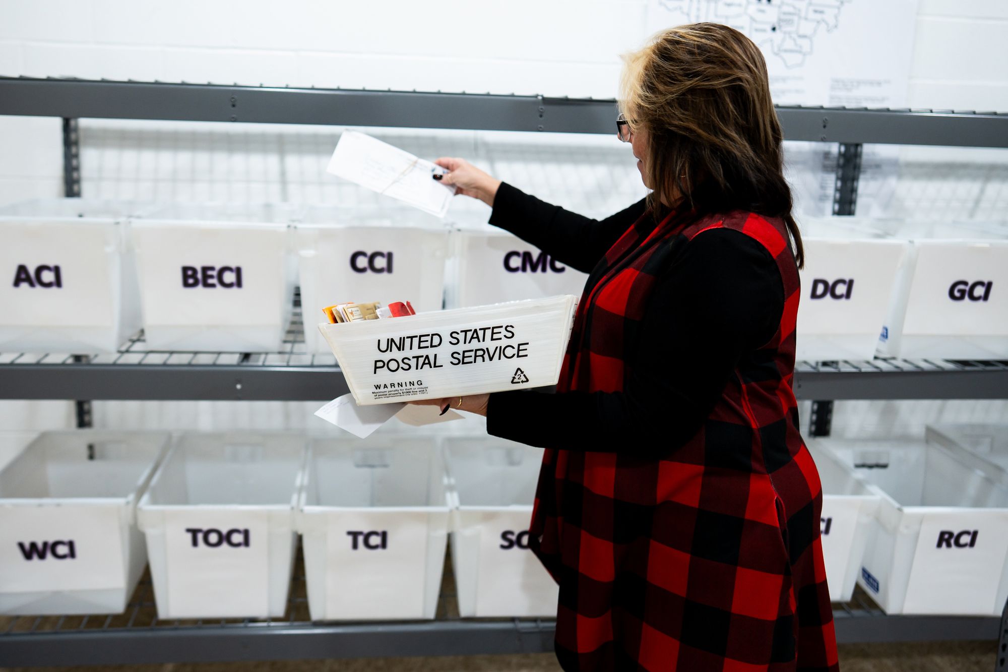 A photo shows Tawnya Gray, a White woman wearing a red and black plaid dress, sorting mail in front of two shelves holding plastic rectangular buckets. 