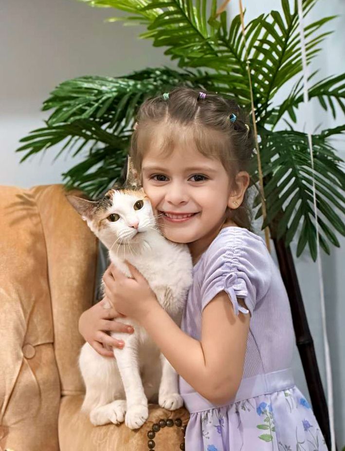 A photo shows a girl with medium-toned skin and dark brown hair hugging a cat with mostly white fur and some brown and black markings on its head. A plant is visible behind them. 
