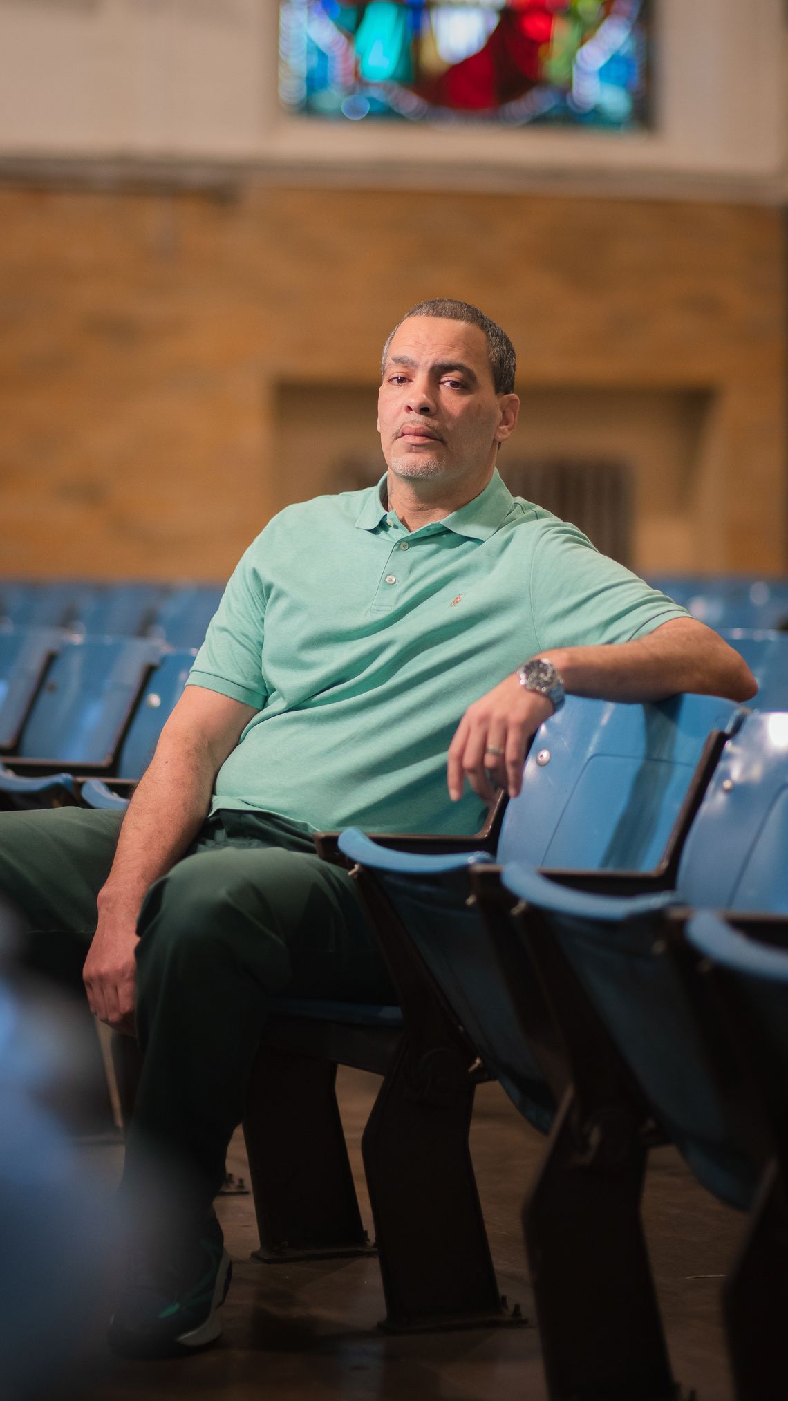 A man with light skin tone and a white goatee, wearing a turquoise polo shirt and green prison pants, sits in a theater chair, his left arm balancing on the seat next to him. 