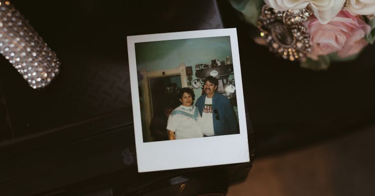 A Polaroid is on a black table. It shows Armando Navejas, a medium light-skinned man with a mustache, standing with his wife Josephine, a medium light-skinned woman. 