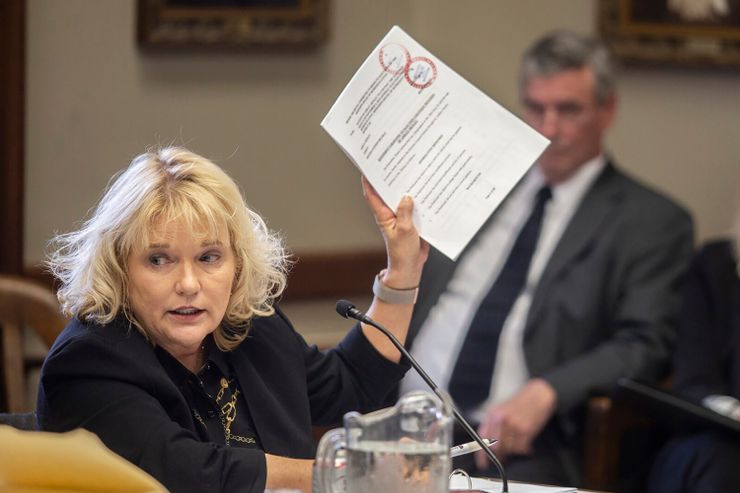 A photo of a White woman with short blonde hair in a black suit, speaking into a mic as she holds up a paper document. Two people are sitting in the background. 