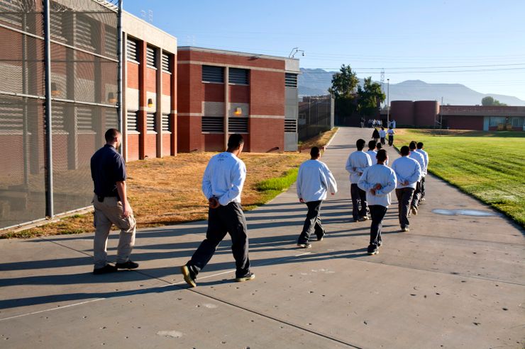 A group of male teenagers dressed in gray sweaters and black pants are walking in two lines, with their hands behind their backs. A man in a dark blue collared shirt and tan cargo pants walks behind them. 