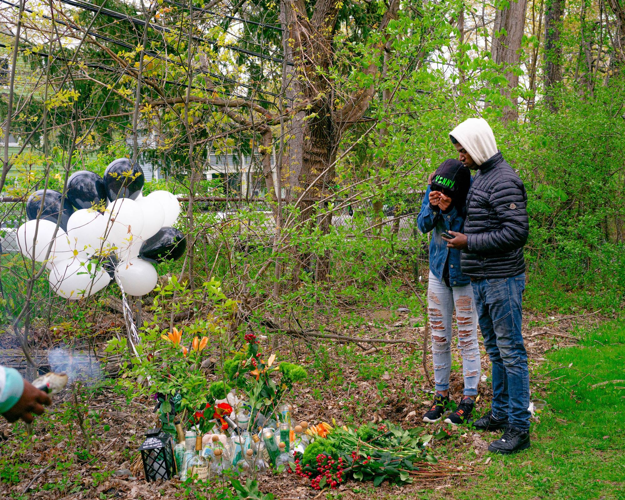 A photo of a man and a woman standing beside memorial flowers, candles, drinks and balloons on the ground. 