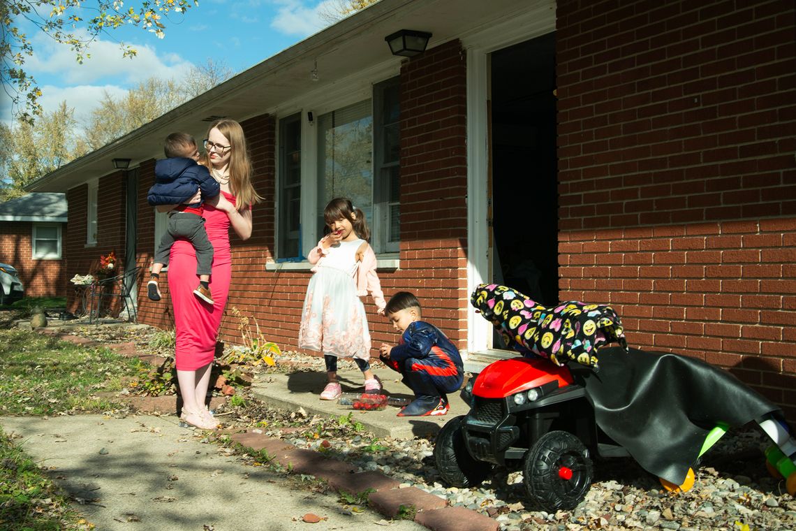 A White woman, wearing glasses and a red dress, holds one of her children as she stands in front of her house. Her other two children, who are standing beside her, are eating strawberries.