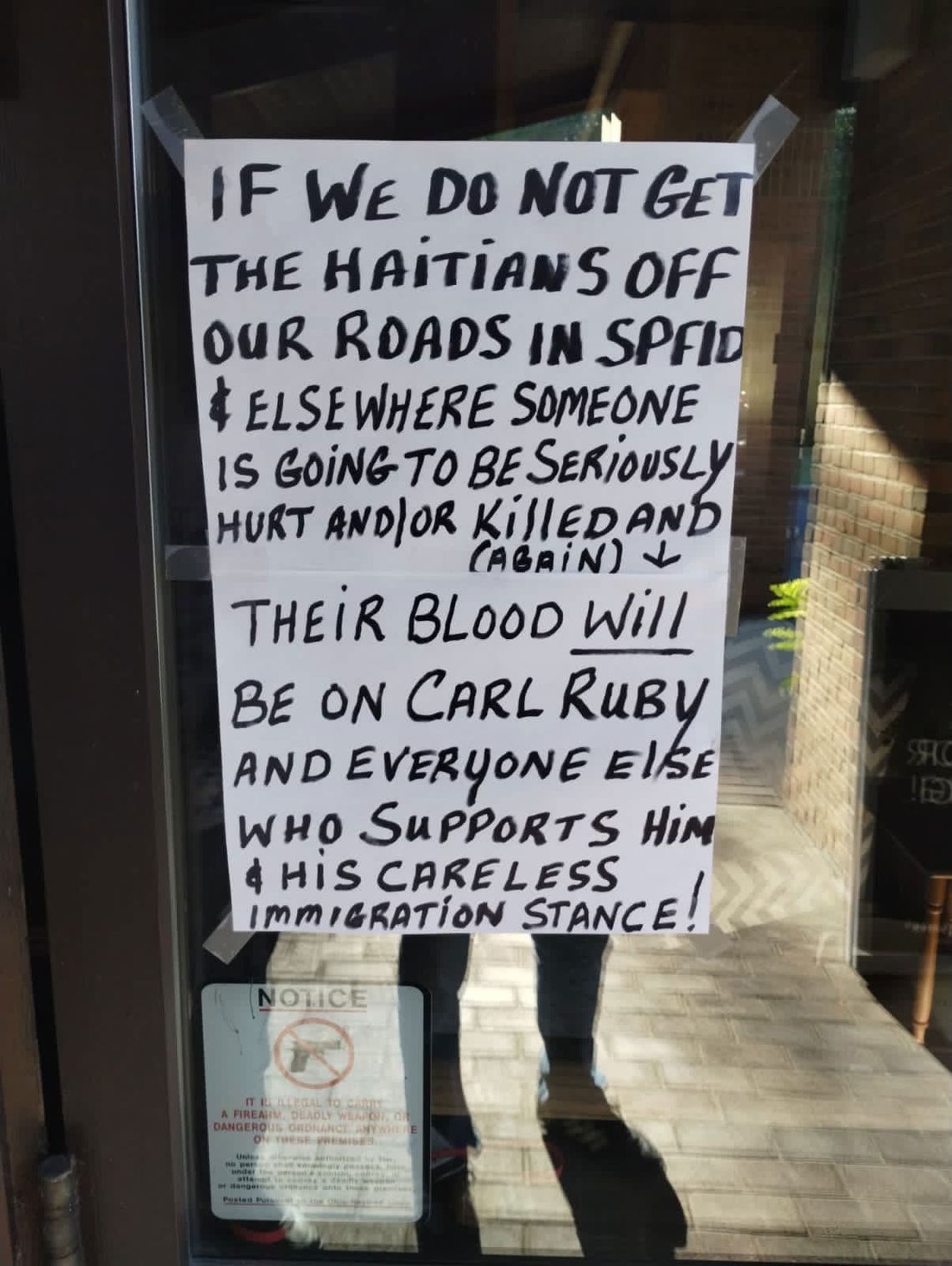 A photo shows a sign written in black marker on white paper, taped on a glass door. The sign reads: “If we do not get the Haitians off our roads in SPFLD & elsewhere someone is going to be seriously hurt and/or killed (again). Their blood will be on Carl Ruby and everyone else who supports him & his careless immigration stance!” 