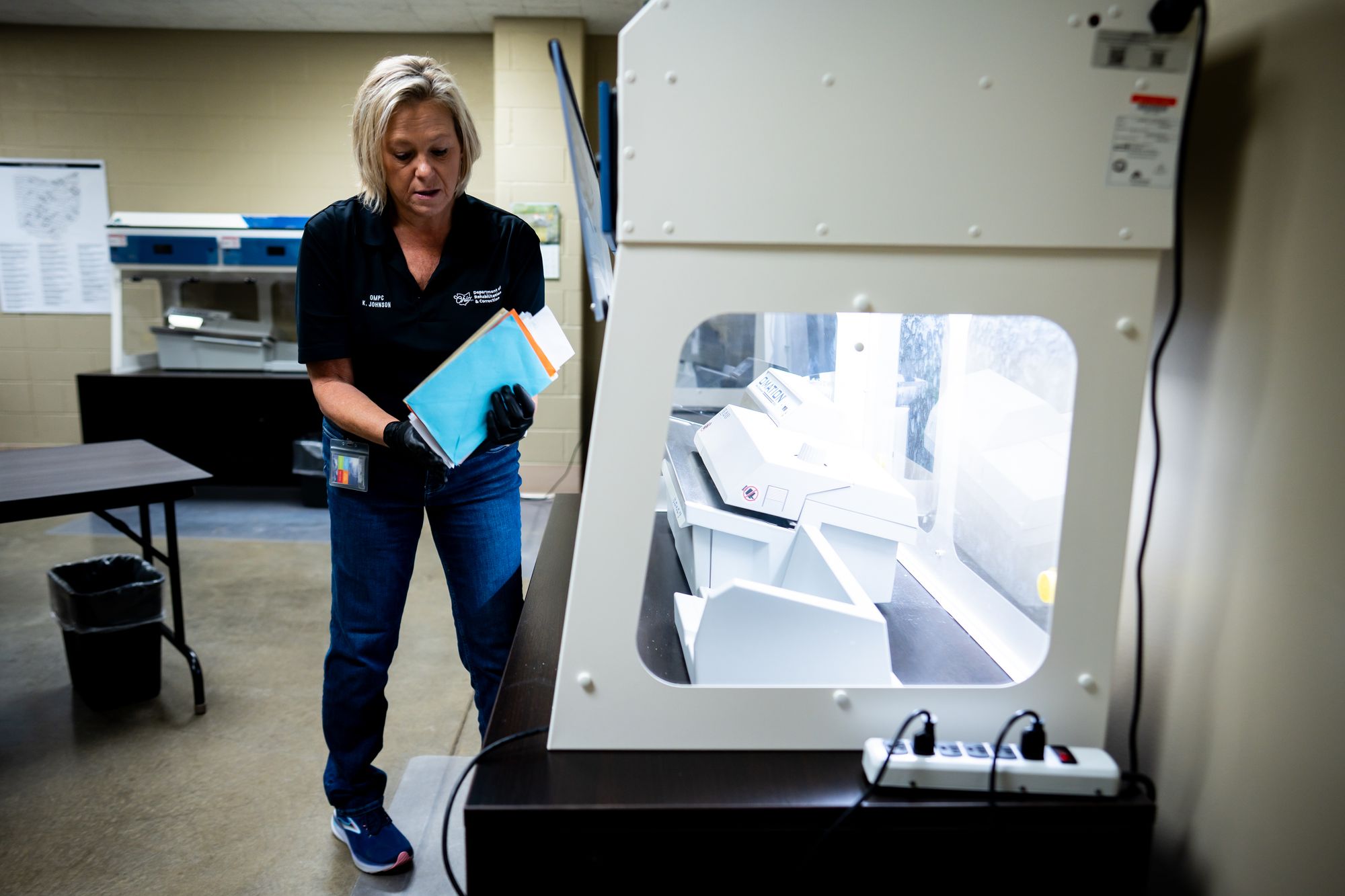 A photo shows Karen Johnson, a White woman, holding some pieces of mail while standing to the left of a machine in a mail processing room. 
