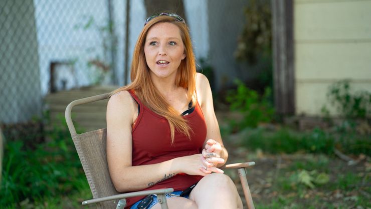 Amanda Aguilar, a woman with light-toned skin and red hair, wears a red tank top and denim shorts while sitting on a lawn chair outside her home.  