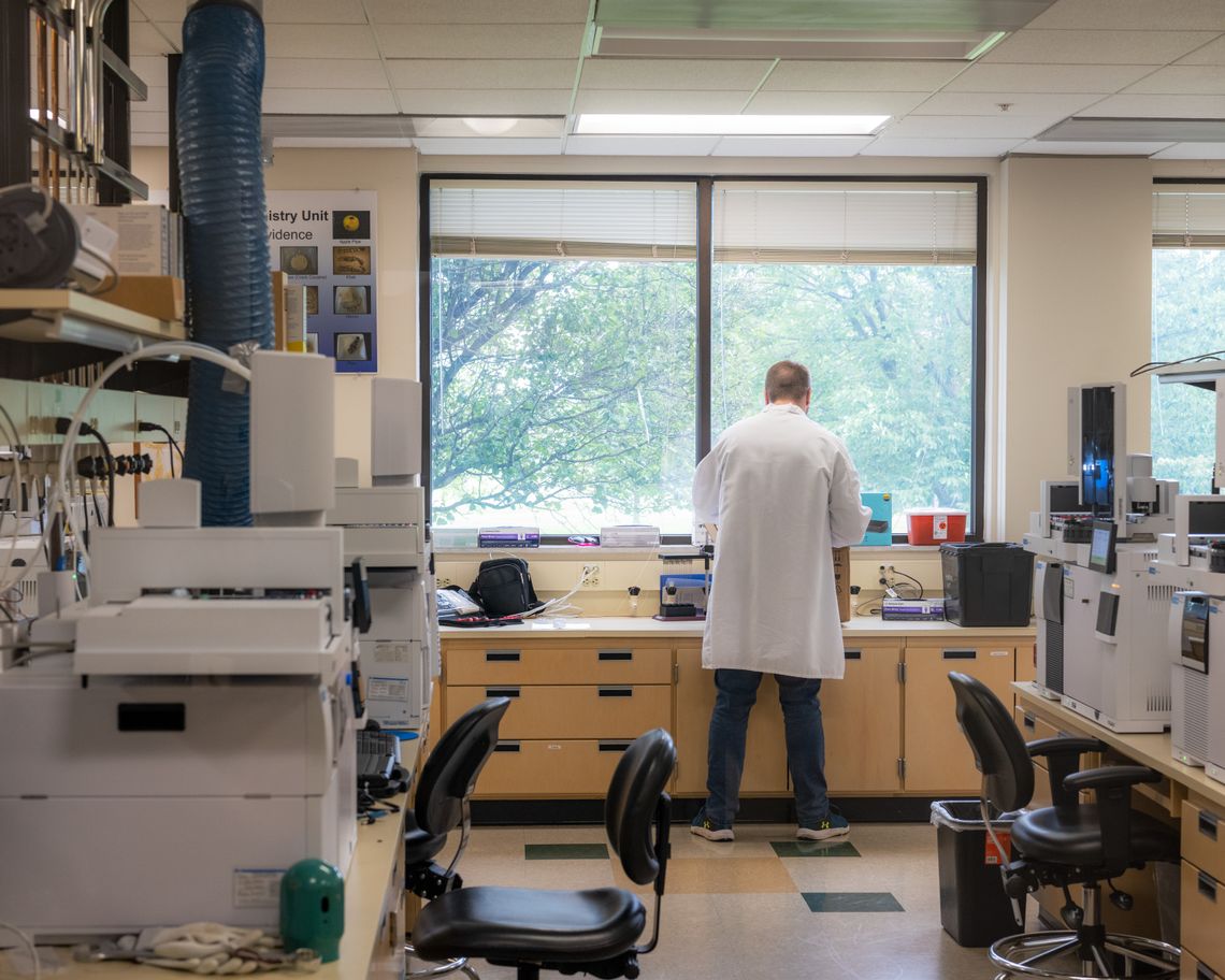 A photo shows a man in a lab coat facing a window in a lab, away from the camera. There are a variety of machines around the lab. 