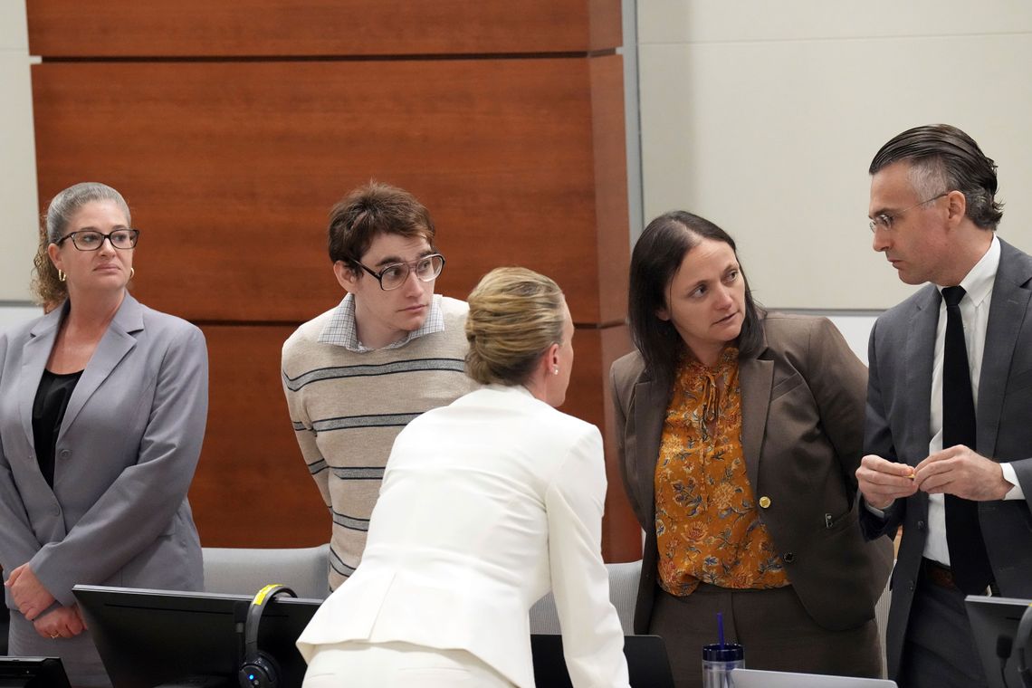 A photo of the defense team and a young White male defendant in a courtroom. Two White female defense team members wearing dark-colored suits are looking toward their colleague, a middle-aged White man with slicked-back dark hair wearing a suit. A third White woman on the defense team has her back to the camera while wearing an all-white suit.