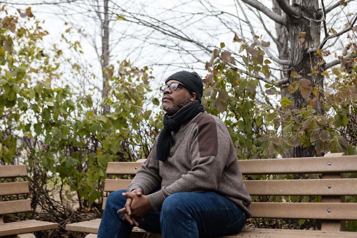 Christopher Thorpes, in a hat and scarf, sits on a bench near some trees. 