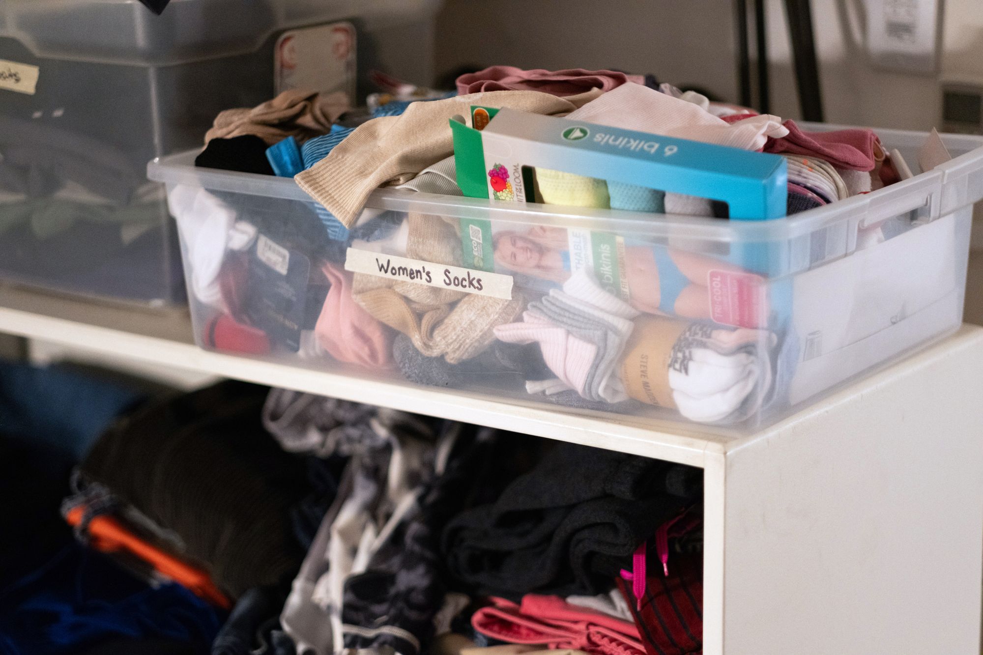 A plastic tub of women's socks sits on a white shelf that holds pairs of pants.