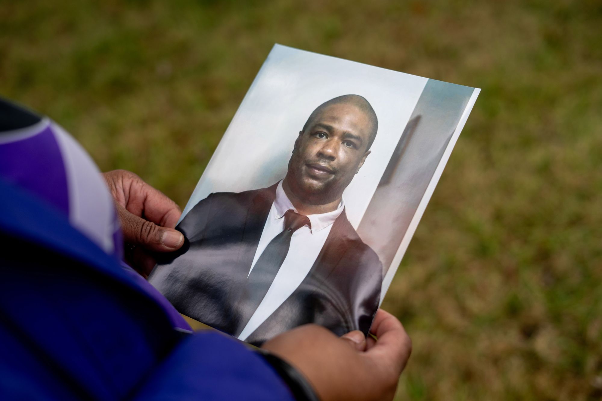 A Black woman’s hands hold a photo of a Black man wearing a black suit jacket, white shirt and black tie.