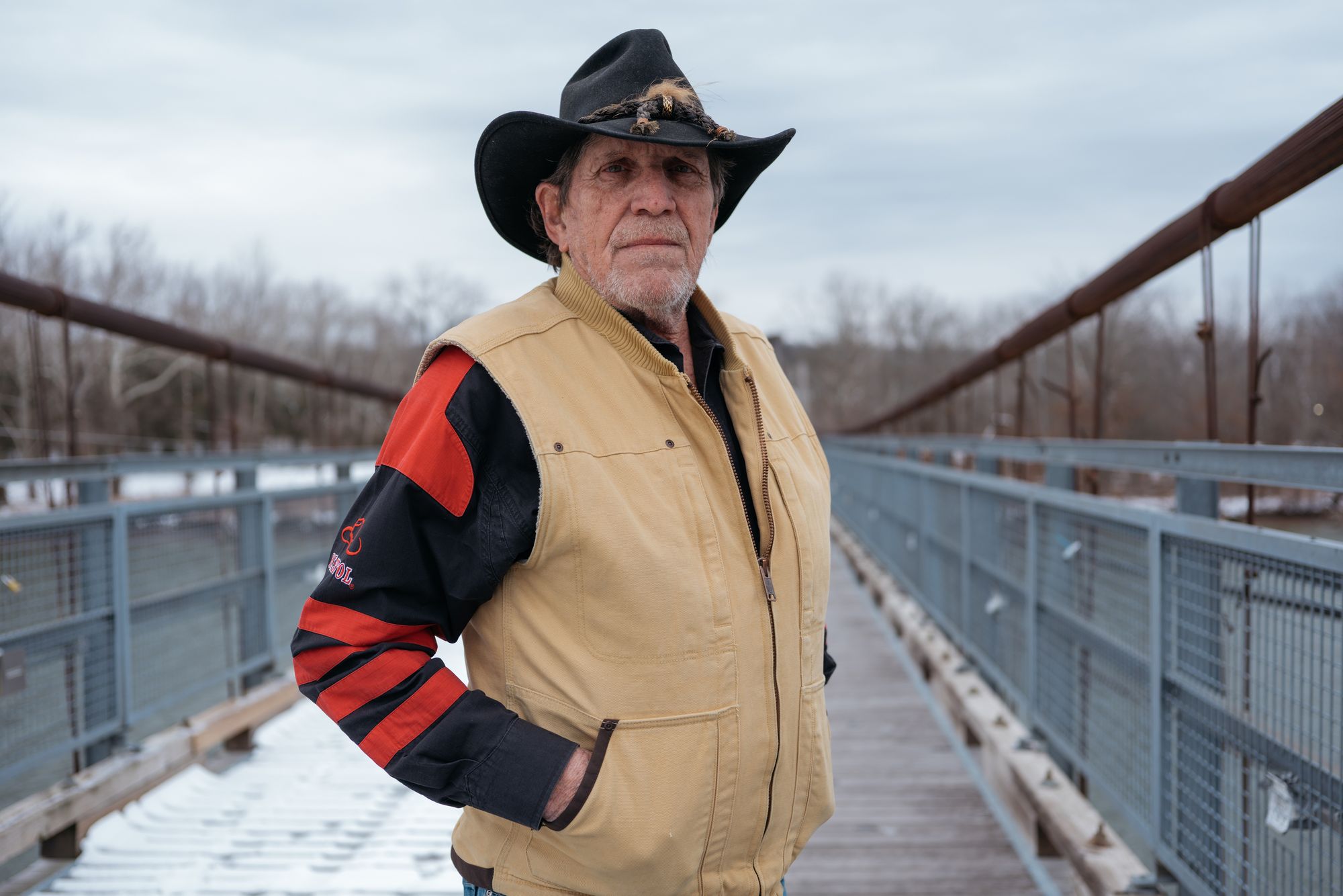 A photo shows a White man standing on a bridge in winter. He is wearing a cowboy hat and a beige vest over a red-and-black jacket. 