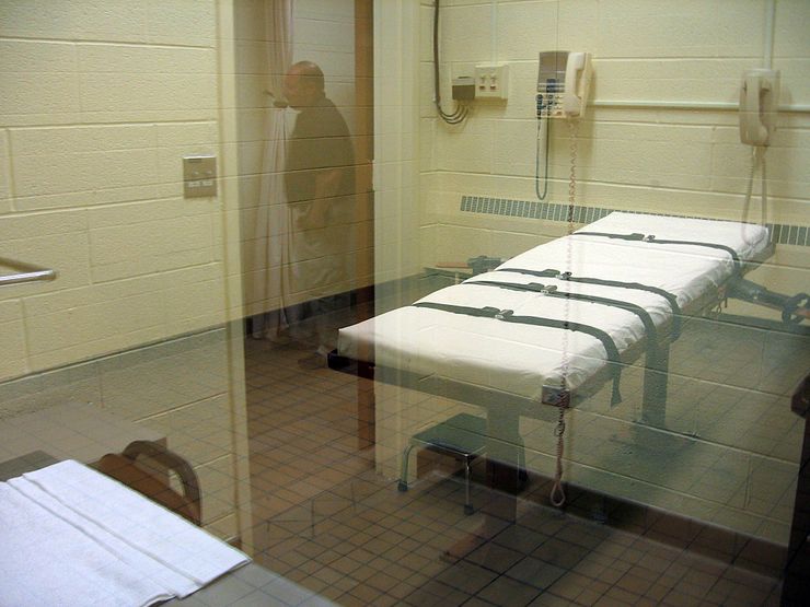 A photo shows a view through a glass panel of a white gurney with black straps in a mostly empty room. On the wall of the room there are two landline telephones. In the reflection of the glass, there is a middle-aged man standing next to a curtain seen through a doorway.  