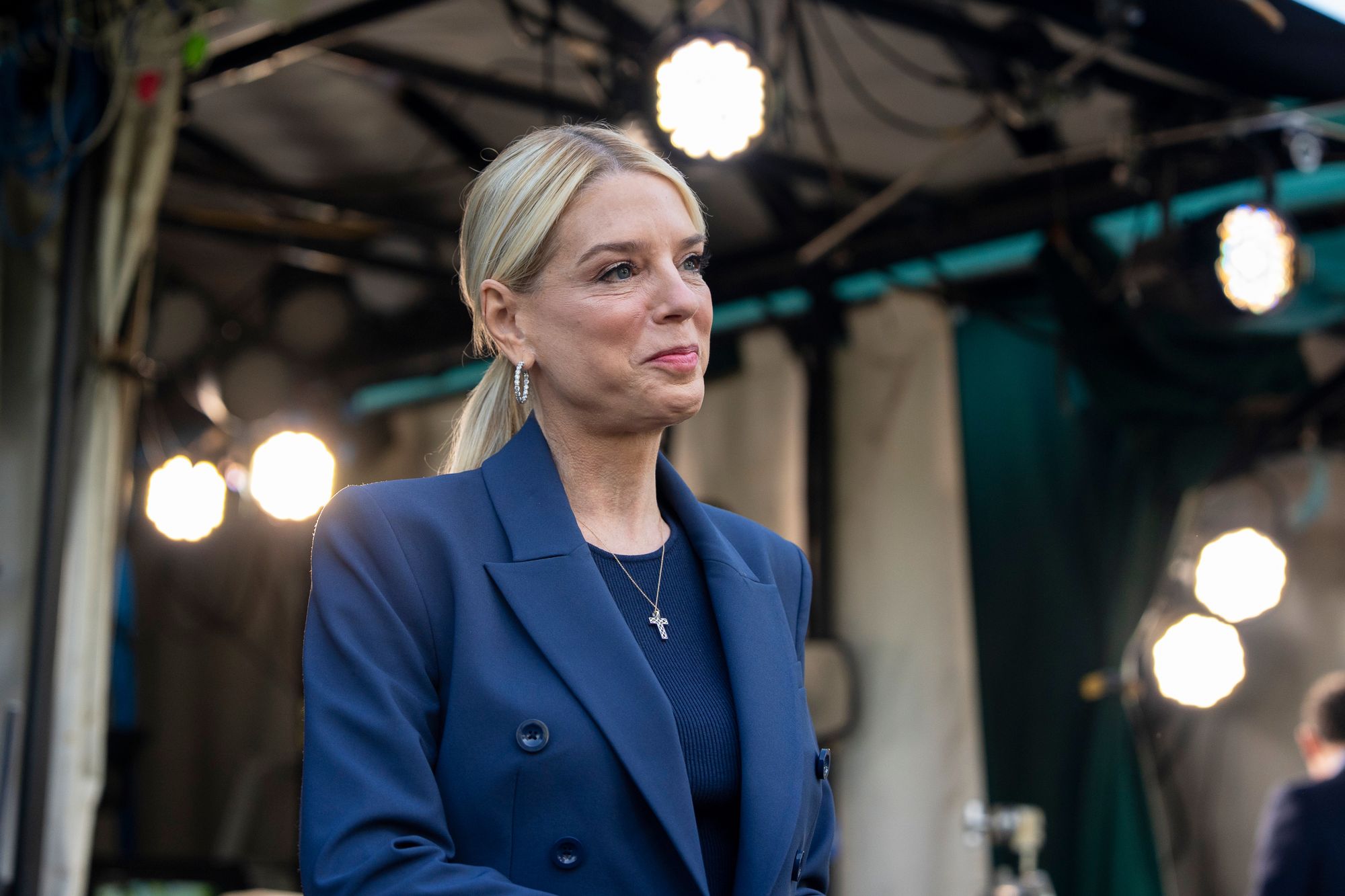 A woman with blonde hair wearing a blue suit walks in front of a tent with overhead lights.
