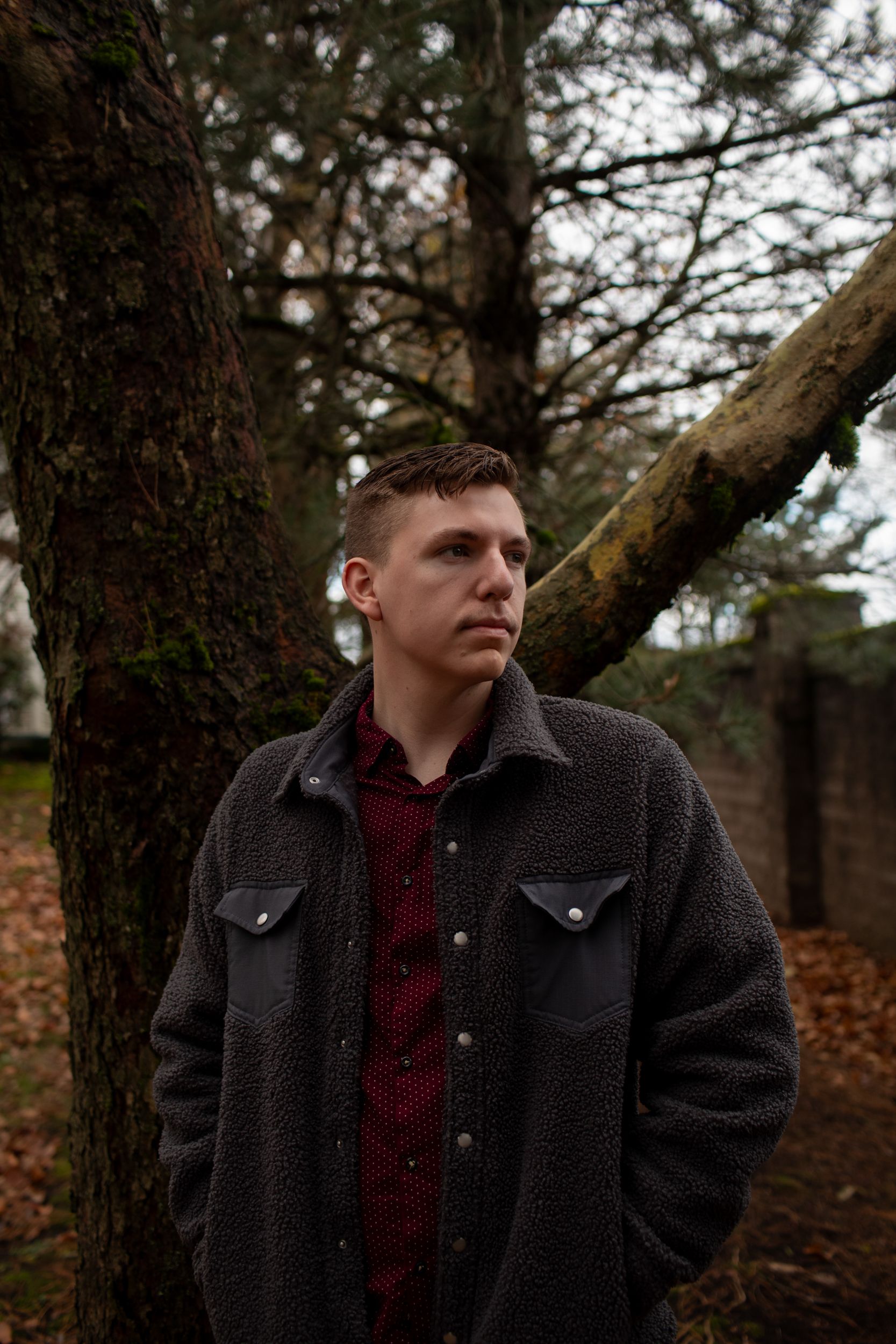 A White man with short brown hair, wearing a fleece jacket, stands in front of a tree on an overcast day, looking off to the side. 