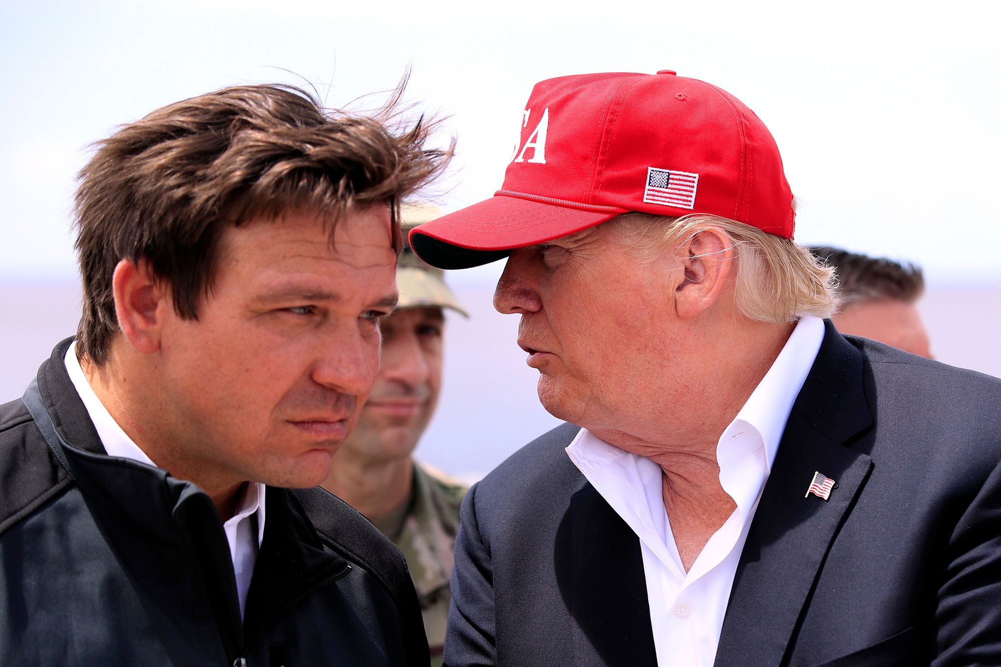 A White man in a black jacket leans his head forward as another White man, who is wearing a red cap and suit, talks to him. 