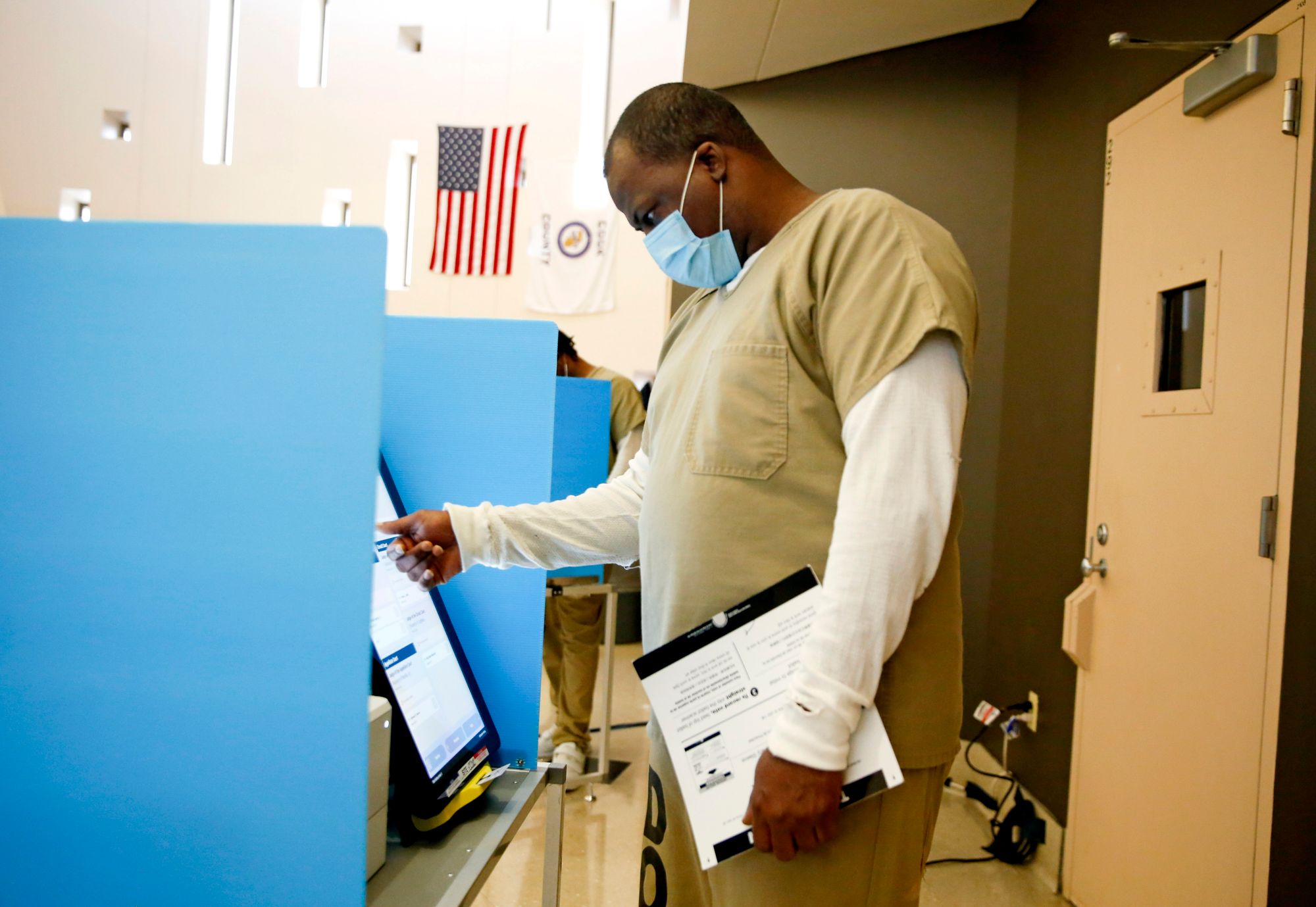 A Black man in a tan prison uniform uses a touch screen to vote at a polling place inside a jail. 