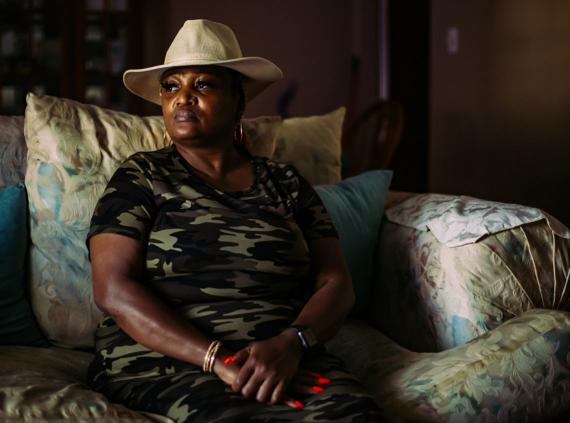 A Black woman looks away from the camera while sitting and wearing a khaki hat and a camo dress.
