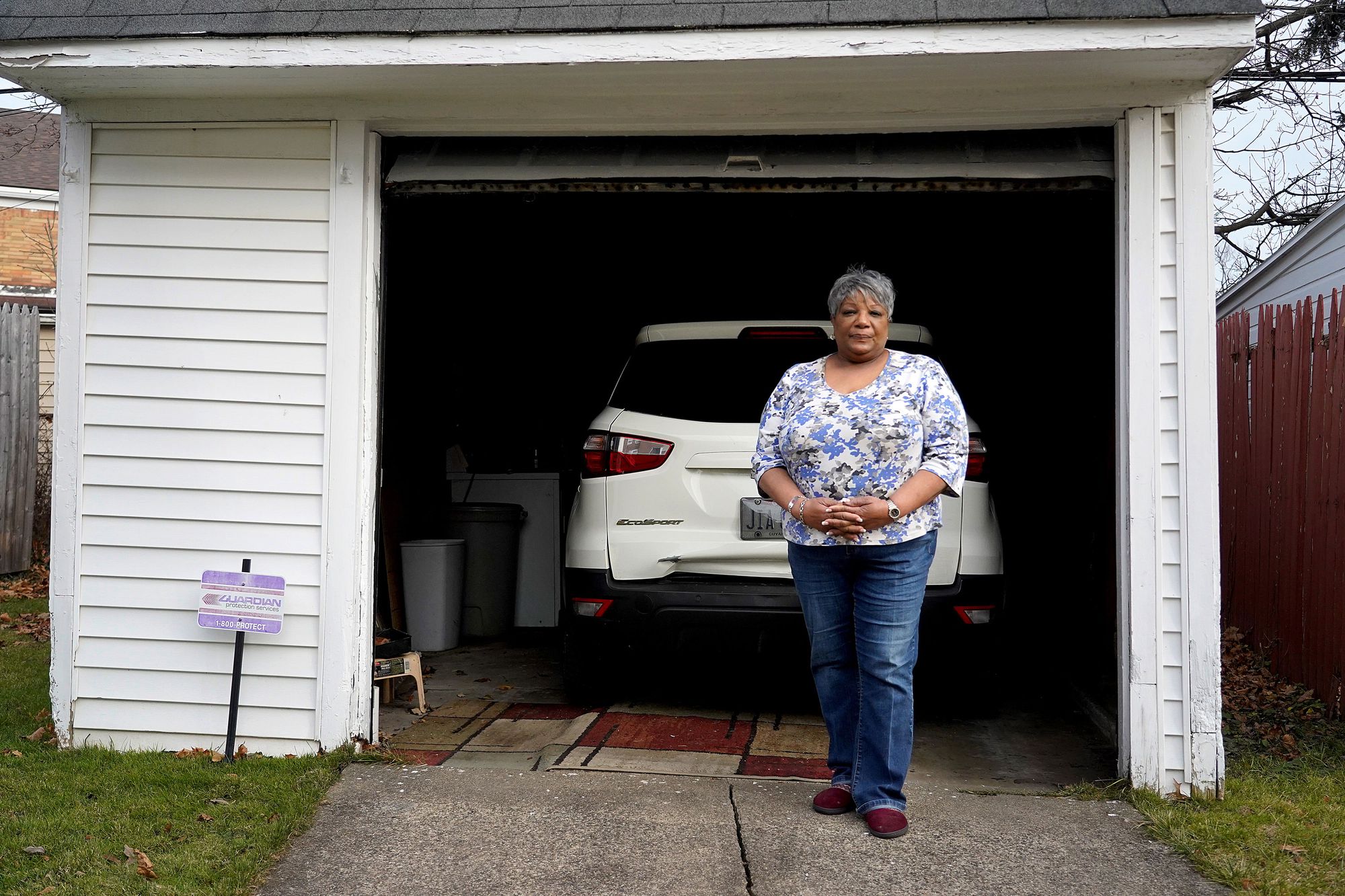 A Black woman with short, gray hair and a blue, patterned top poses for a portrait in front of her white car in a garage. 