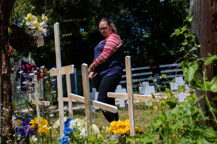 A woman wearing glasses and with long brown hair in a ponytail stands in front of wooden crosses and flowers. 