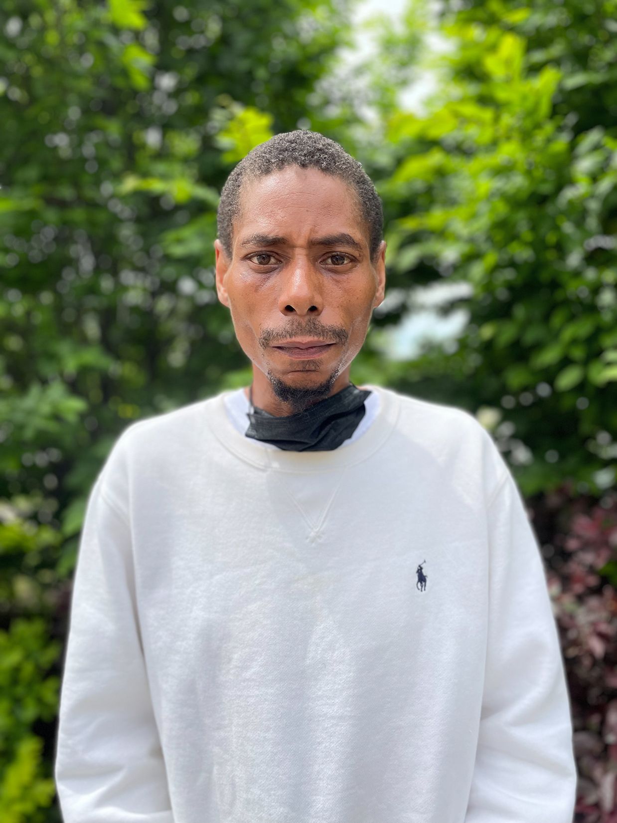 Harold Scott, a Black man with short hair, a thin mustache, and goatee, wears a white sweatshirt against the backdrop of green leaves on trees. 