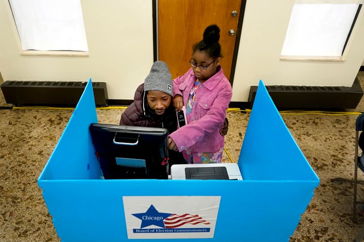 A Black man, wearing a gray beanie and brown puffer jacket, sits in front of a touch screen monitor at a voting booth. He looks at the screen together with his daughter, who is wearing glasses, a floral shirt and pink jean jacket.