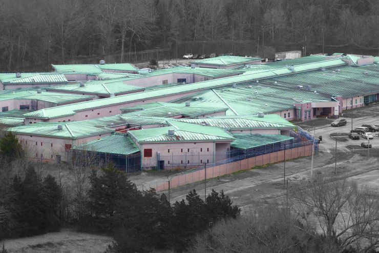 An aerial photo of a prison facility with a light teal roof and its adjacent parking lot. Bare trees surround the facility. 