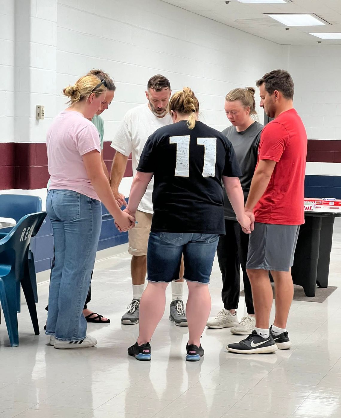 A group of six people hold hands with their heads facing downward. They are standing in a circle, inside a prison visitation room.