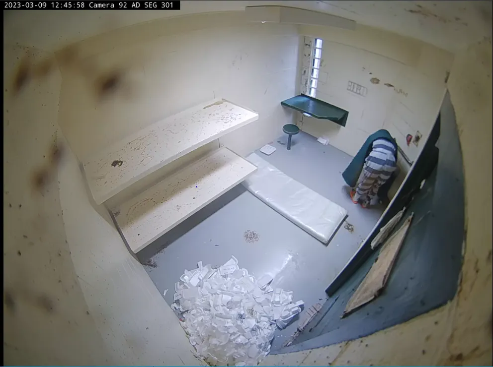 A still from a video shows a man, wearing a gray striped uniform, leaning toward a sink in the corner of his jail cell. A large pile of white containers fills a different corner of the cell.