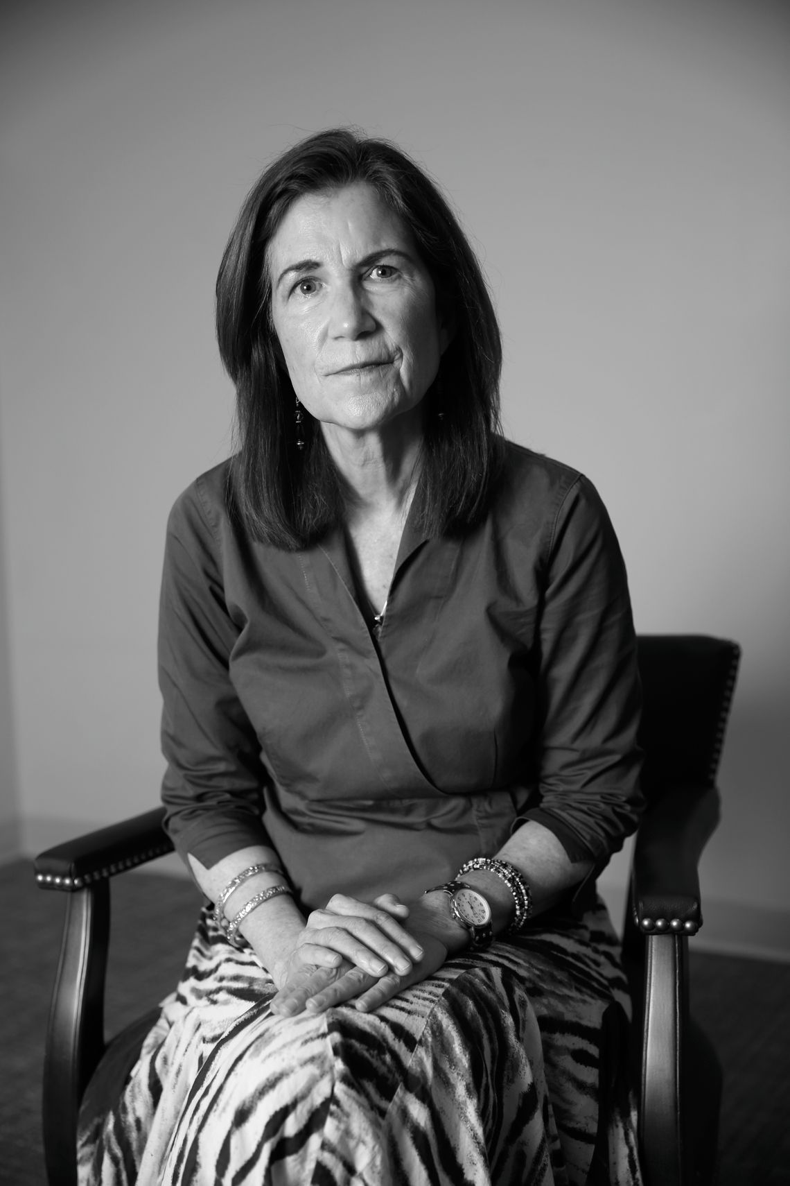 A White woman faces the camera for a portrait shot. She sits on a chair, with her hands placed on top of each other on her lap.