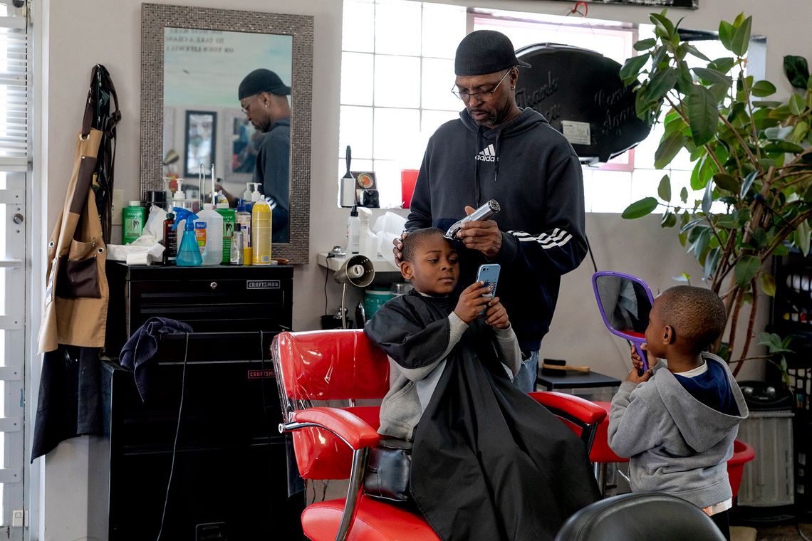 A Black man wearing a black sweater, glasses and hat, uses a clipper to cut a Black boy’s hair inside a barbershop. Another Black boy, wearing a gray sweater, stands next to the barber’s chair while holding a mirror. 