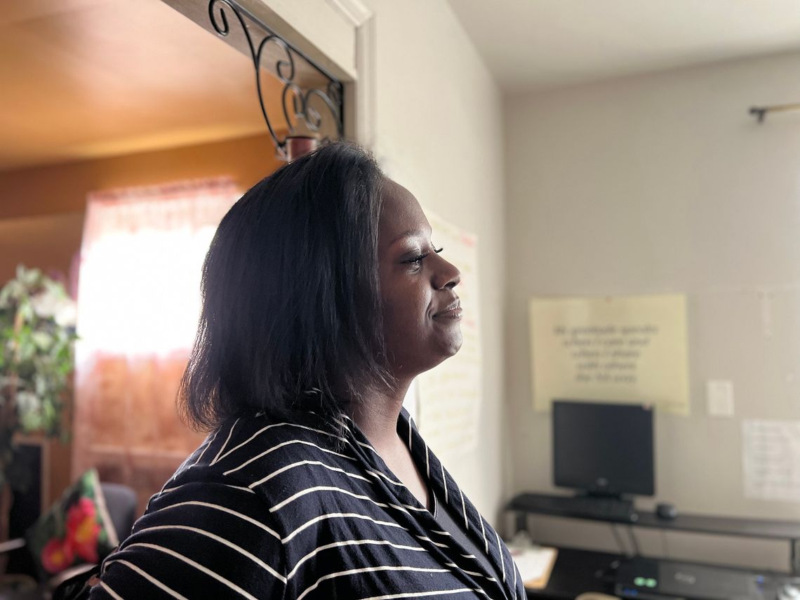 A Black woman wearing a black-and-white striped shirt stands in a room. A computer, a desk and a poster are in the background. 