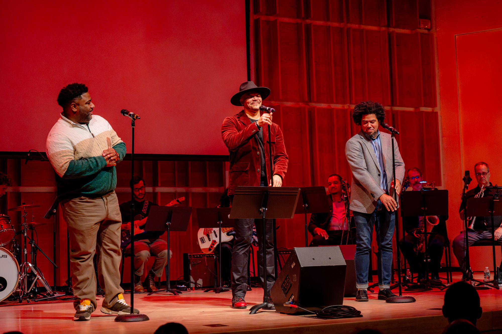 Three men stand on a stage behind microphones, in front of a row of seated people playing musical instruments, including a guitar, an electric bass, trombone, saxophone and trumpet. At left is a Black man with a short beard, wearing tan pants and a quarter-zip sweater that is white, cream and green. In the center, a Black man wearing red shoes, black pants, a red velvet jacket and a wide-brimmed black hat. At right stands a Black man with a goatee wearing jeans, black shoes and a gray sport jacket.