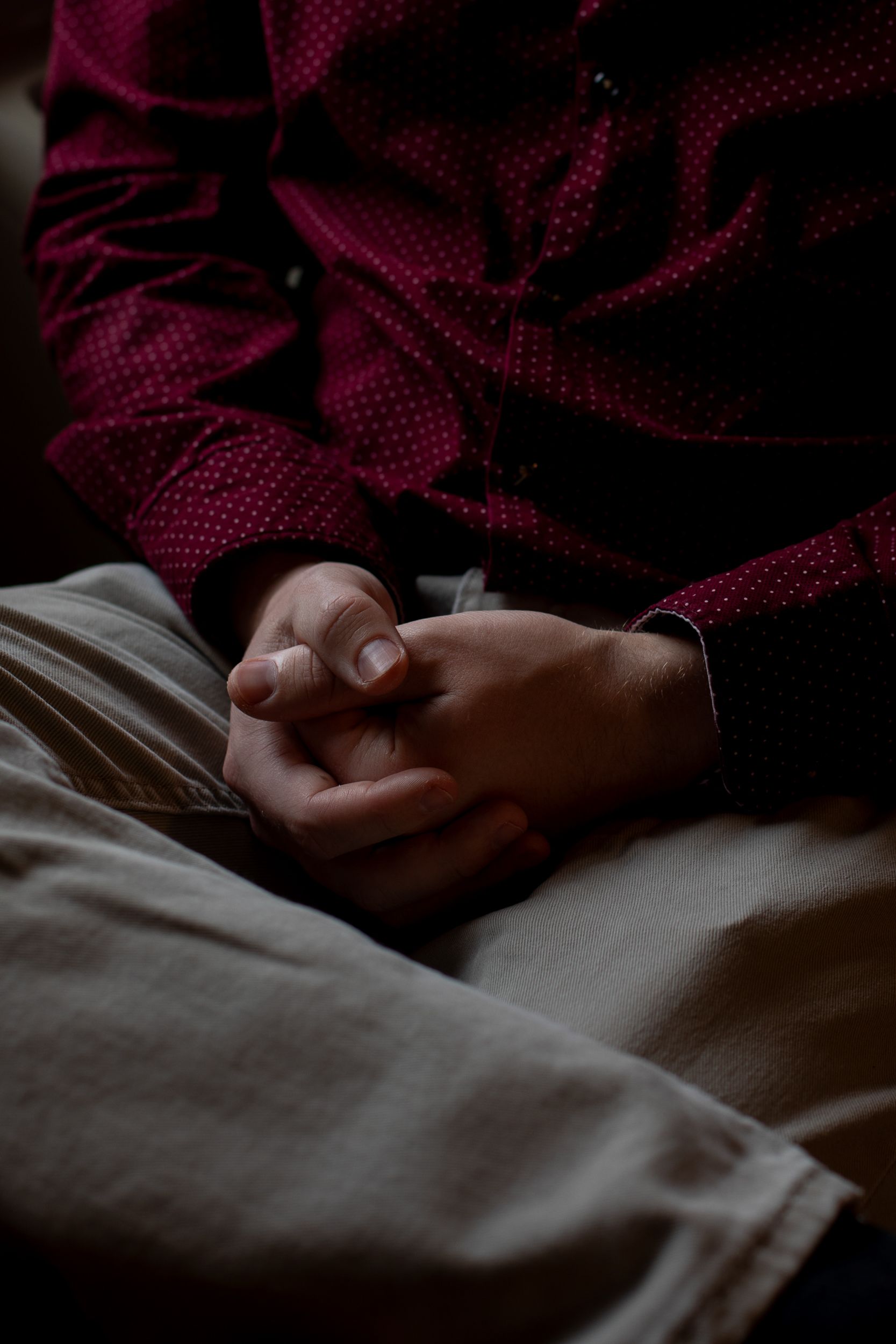 A close-up of light skin-toned hands, resting on the lap of a person wearing beige pants and a maroon shirt. 