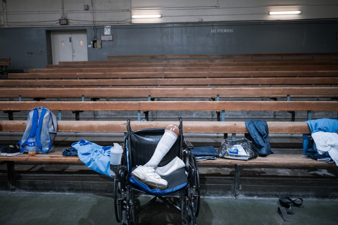A photo shows the bleachers of a gym. In front of the bleachers is a wheelchair with a prosthetic leg in it. 