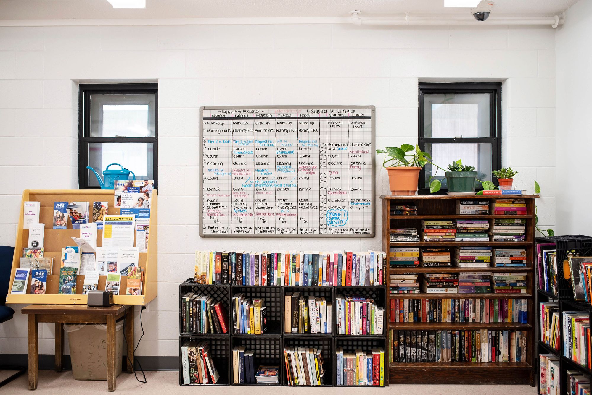 A room with two small windows has two bookshelves situated against the wall, filled with books. One of the bookshelves has potted plants on the top row. There is a display shelf with brochures on it to the left. A white board hangs in the center of the wall, with a schedule of activities written on it, including “Wake up”, “Morning Circle”, “Lunch”, “Count”, “Cleaning” and “Free Rec”. 