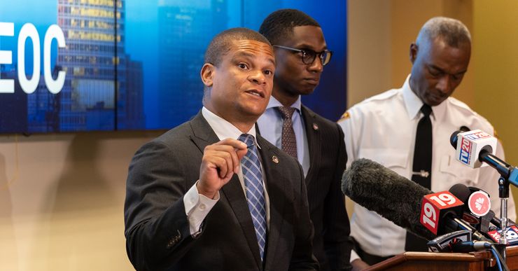 A Black man in a dark gray suit stands at a podium during a press conference. 