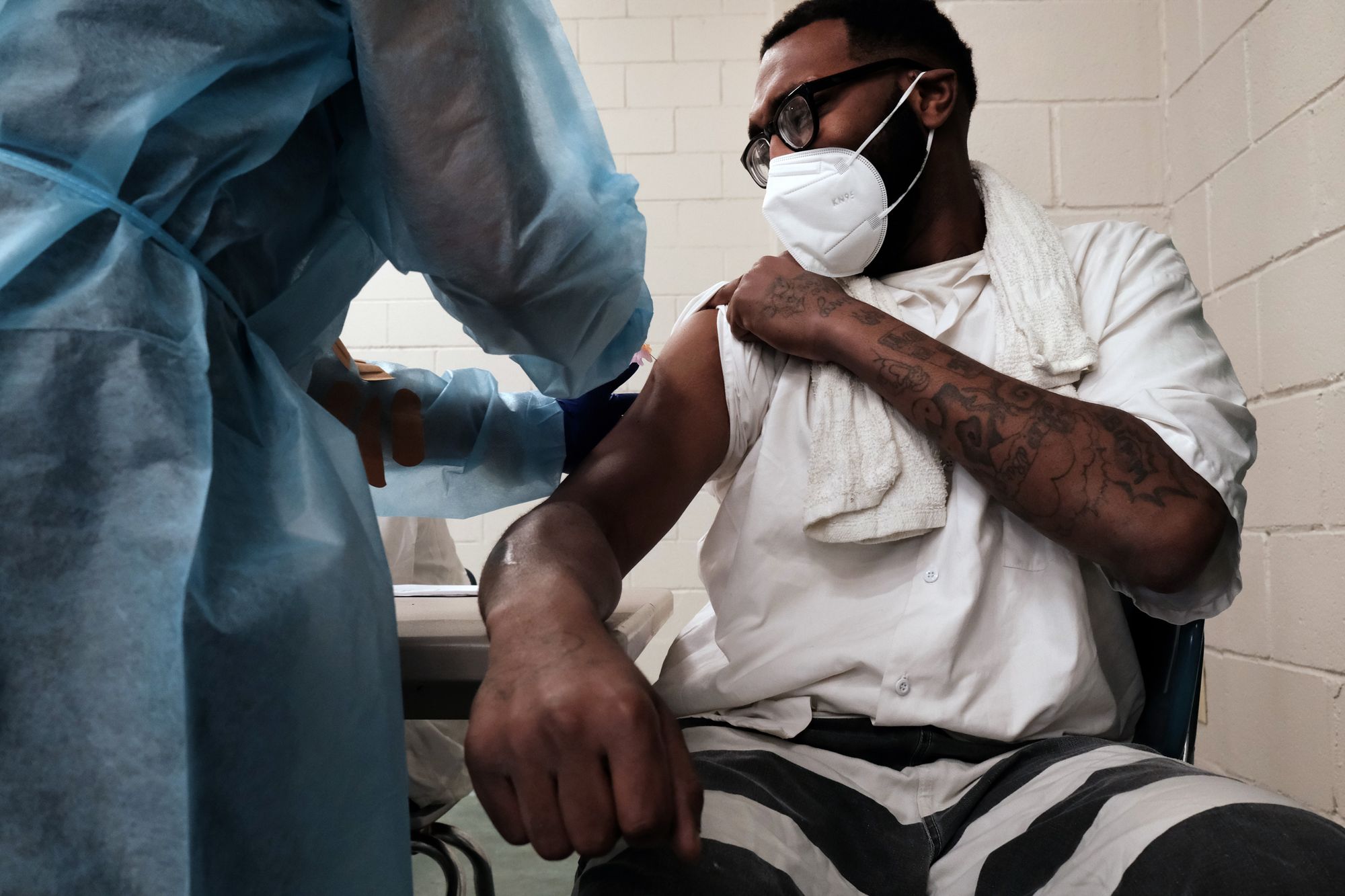 A man with glasses in a prison uniform receives a vaccination from a person in a blue lab coat.  