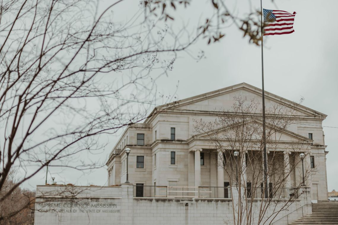 A color photo shows a white stone building with columns and steps leading up to it, and an American flag on a pole. The side of the building has sign with block lettering that reads “Supreme Court of Mississippi” and “Court of Appeals of the State of Mississippi.”