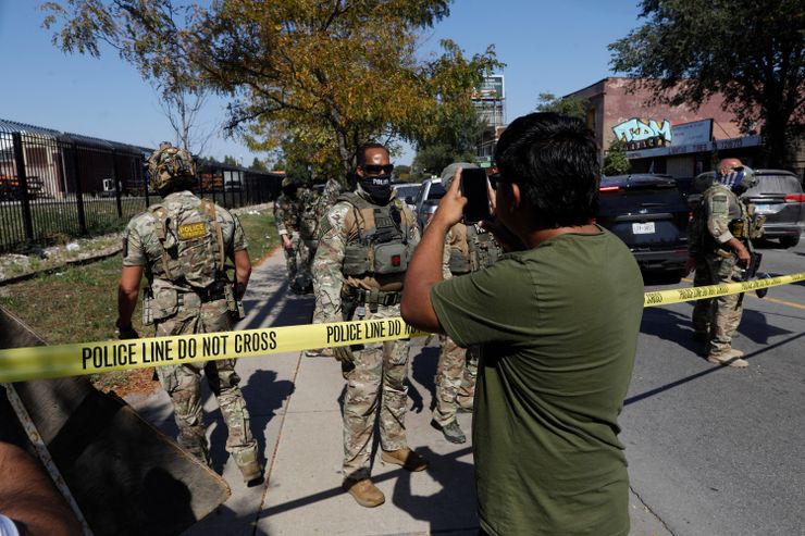A photo shows a man with a medium skin tone and a olive green t-shirt holding his phone up toward a group law enforcement agents, who are standing in uniform on the other side of yellow police tape. 
